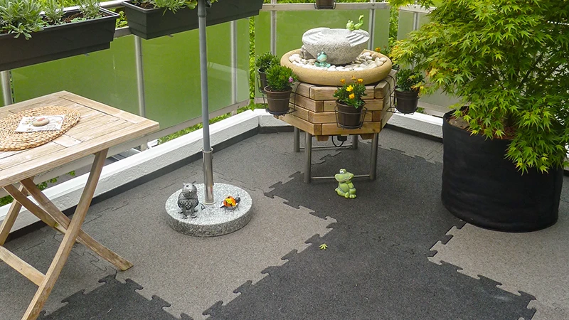 Cette photo montre un balcon accueillant, recouvert de dalles puzzle WARCO bicolores. Une petite fontaine, une étagère en bois et plusieurs jardinières créent une atmosphère conviviale. Le sol offre un grand confort grâce à sa surface élastique et antidérapante, et il est également résistant aux intempéries. Grâce à l’assemblage précis, on peut ajouter des touches de couleur qui donnent une touche moderne à l’espace extérieur.