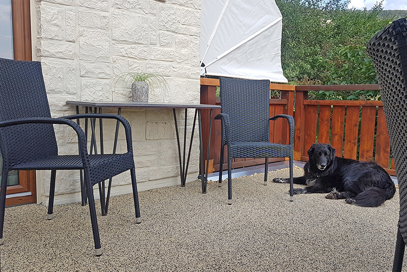 Schwarzer Hund liegt auf Balkon mit grauen  Balkonplatten. Zwei Stühle und ein Tisch stehen am Haus. Über ein braunes Holzgeländer geht der Blick auf grüne Bäume.