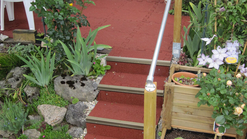 rote Treppe führt auf Terrasse. Rund um den WARCO-Treppenbelag wachsen grüne Pflanzen.