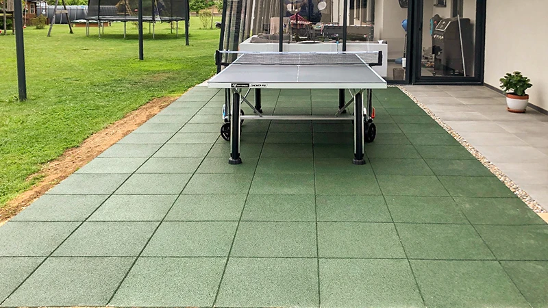 Auf einer grünen Terrasse aus WARCO-Platten aus pu-gebundenem Gummigranulat steht eine Tischtennisplatte vor einem luxuriösen Haus. Im Garten sind ein Trampolin und weiteres Spielgerät platziert, was zu aktiver Erholung und Bewegung im Freien einlädt. Die robusten, rutschfesten Terrassenplatten sorgen für Komfort und Sicherheit. Ihre elastische Oberfläche schont die Gelenke, während die pflegeleichte Struktur einen sauberen Look bewahrt.