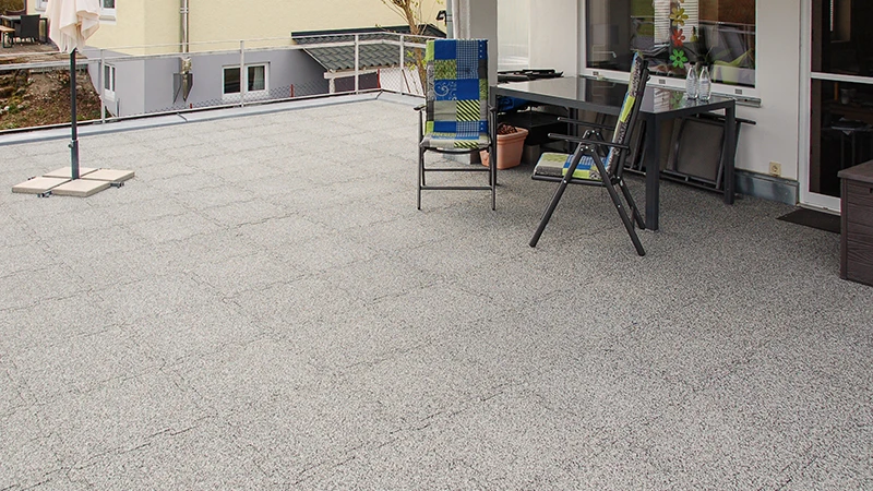 Sur un toit-terrasse sont installées des dalles de terrasse WARCO gris marbré en granulat de caoutchouc lié au PU. Leur emboîtement puzzle assure un ajustement précis, alliant résistance aux intempéries et durabilité. Le matériau, stable dans le temps, atténue les bruits de pas et renforce l’aspect net et soigné de la surface. Il procure également une bonne adhérence et un confort de marche élevé, rendant le revêtement très fonctionnel.