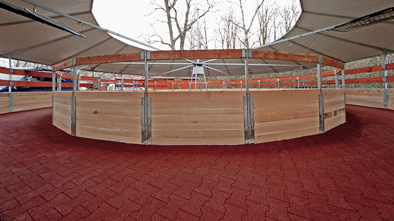 Das Foto zeigt eine moderne, überdachte Führanlage für Pferde. Der Hufschlag ist durchgehend mit rotem Gummi-Verbundpflaster belegt – rutschhemmend, wasserdurchlässig und besonders gelenkschonend. Die elastische Oberfläche ermöglicht sicheres Bewegen der Pferde bei jeder Witterung und in jeder Gangart und reduziert die Belastung auf Hufe, Gelenke und Sehnen. Der befestigte Hufschlag ist formstabil, langlebig und besonders pflegeleicht. Die Anlage eignet sich ideal für Reitbetriebe, Zuchtställe und Pferdekliniken, die auf tiergerechte Bodenverhältnisse, minimalen Pflegeaufwand und dauerhaft belastbare Materialien setzen.