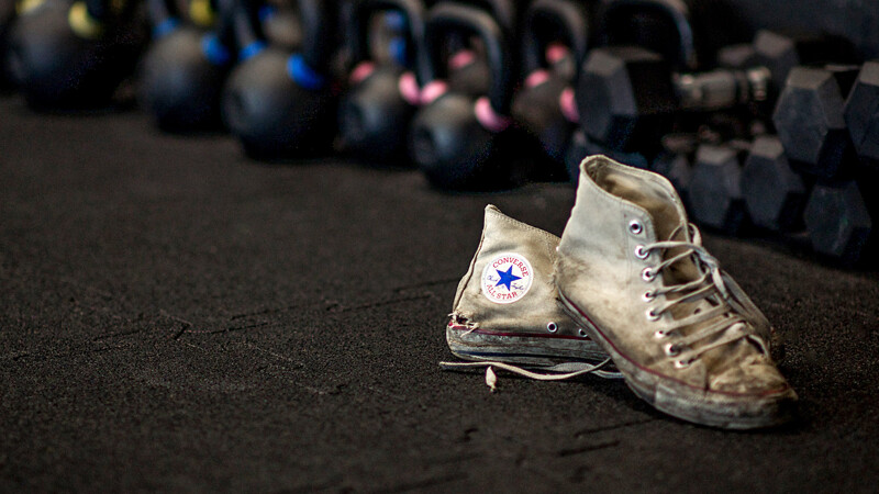 Ein Paar stark gebrauchter Converse ALL STAR Sneakers steht vor einer Reihe von Hanteln im Fitnessstudio auf schwarzen Fitnessmatten von WARCO.