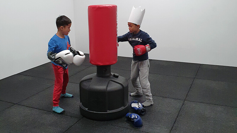 2 Kinder boxen auf Boxsack, schwarze Fitnessmatte von WARCO bieten guten Fallschutz und Halt.