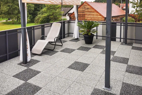 Die sanierte Dachterrasse ist mit Terrassenplatten in Granitoptik gestaltet. Die Platten im geometrischen Muster aus hellgrauen und dunkelgrauen Quadraten schaffen eine moderne und stilvolle Atmosphäre. Der robuste Bodenbelag aus Gummiplatten bietet Trittsicherheit und Komfort. Ein Sonnenschutz spendet Schatten an heißen Tagen. Ein Pflanzkübel mit Palme und eine Liege ergänzen die einladende Gestaltung der Terrasse. Die Geländerkonstruktion aus Metall und Glas verleiht der Dachterrasse eine offene und luftige Optik.