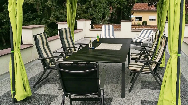 Auf einer Dachterrasse liegt ein Bodenbelag aus WARCO Terrassenplatten in einer hell- und dunkelgrauen Marmorierung, die an Granit erinnert. Ein Baldachin überspannt die Sitzgruppe auf dem elastischen, witterungsbeständigen Belag. Die rutschfesten, stoßdämpfenden Platten lassen sich ohne feste Verklebung verlegen und dämmen Trittschall. Es entsteht eine pflegeleichte Fläche für entspannte Stunden im Freien.