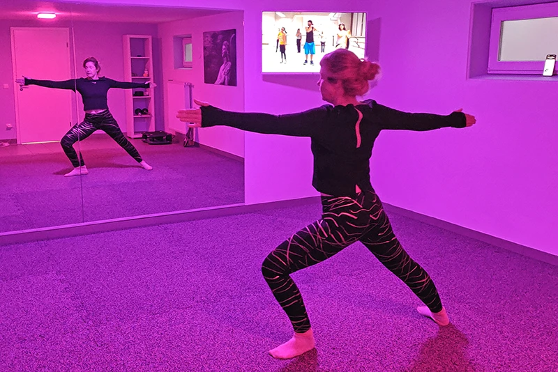 In einem modern eingerichteten Homegym sieht man eine Person in sportlicher Kleidung, die vor einer Spiegelwand eine Yoga- oder Dehnübung ausführt. Der Boden besteht aus hochwertigem, rutschfestem WARCO Fitnessboden, der aus robusten Gummigranulatplatten zusammengesetzt ist. Im Hintergrund ist ein Fernseher an der Wand montiert, der eine Fitnessgruppe zeigt, die als Unterstützung für das Training dient. Die Spiegelwand hilft der trainierenden Person, ihre Übungstechnik zu überprüfen. Der Raum ist mit einer angenehmen Beleuchtung ausgestattet, die eine motivierende und angenehme Trainingsatmosphäre schafft.