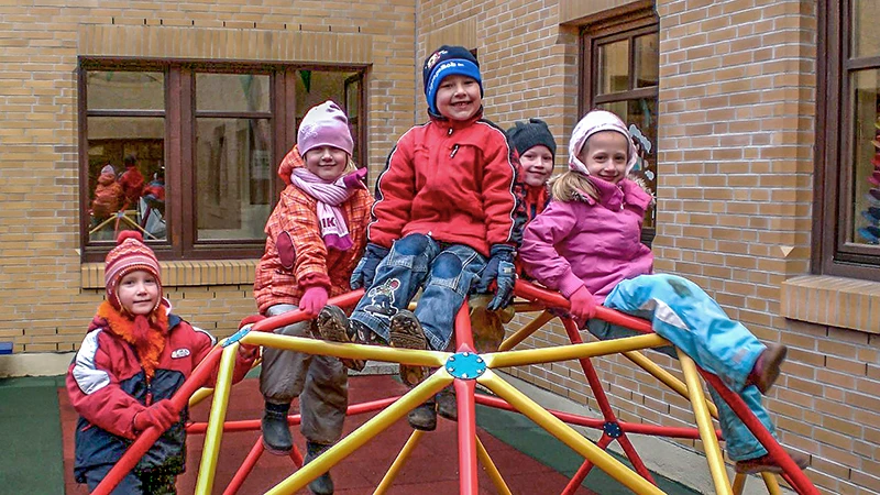 Eine Gruppe von fünf Kindern, warm eingepackt in Winterkleidung, vergnügt sich auf einem bunten Klettergerüst auf einem Spielplatz im Freien. Das Klettergerüst ist ein kleines, kuppelförmiges Klettergerüst aus gelben, roten und blauen Stangen. Die Kinder, die alle fröhlich lächeln, befinden sich an verschiedenen Stellen des Gerüsts: Zwei sitzen oben, zwei stehen und eines hängt an der Seite. Der Boden unter dem Klettergerüst ist mit WARCO Fallschutzmatten ausgelegt, die eine sichere Spielfläche bieten. Im Hintergrund ist ein Backsteingebäude mit großen Fenstern zu sehen, das eine sichere und einladende Umgebung widerspiegelt.