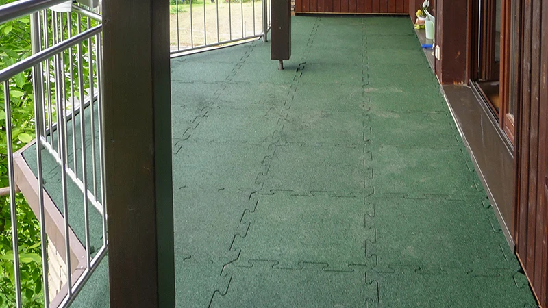 La photo présente un balcon allongé recouvert de dalles WARCO vertes à emboîtement. Leur surface antidérapante et élastique assure sécurité et confort, même en cas d’humidité. Grâce au système d’assemblage précis, on obtient une solution durable et facile à entretenir qui protège le support. Le résultat est un espace extérieur convivial, idéal pour se détendre et profiter de l’air frais.