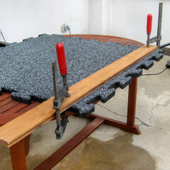 Auf einem Gartentisch mit zwei Schraubzwingen und einem Brett festgeklemmt, lässt sich eine Fallschutzplatte sicher und genau schneiden.