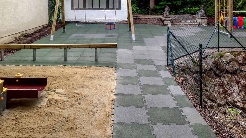 Un terrain de jeu dans le jardin enfants muni de la balançoire et le sol de dalles de jeu de ballon WARCO vertes et grises.