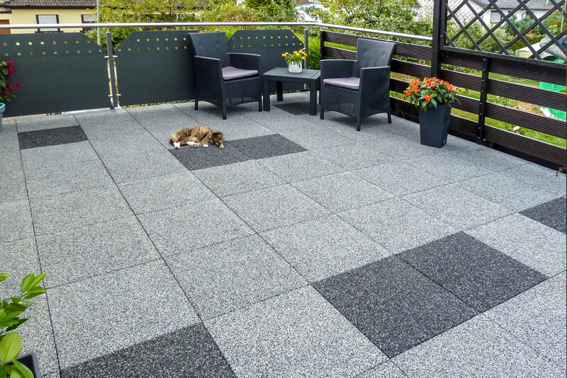 Quadratische Terrassenplatten im Farbdesign Grauer und Dunkelgrauer Granit im Format 50 × 50 cm sind als Boden auf einer Dachterrasse, die sich auf einem Gebäude oder einer Garage befindet, ausgelegt worden. In einer Ecke vor dem Geländer befindet sich eine Sitzgruppe, vor der ein Hund entspannt döst.