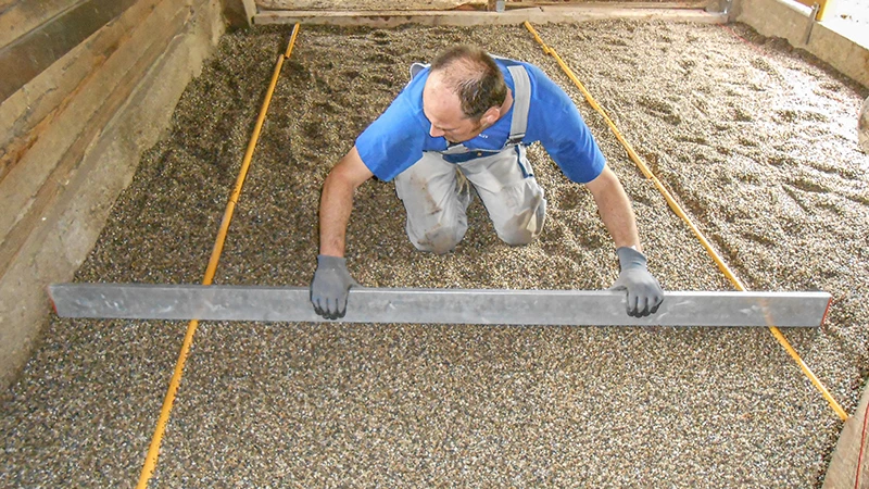 Einbau eines neuen Bodens im Pferdestall: Ein Mann zieht mit einer Richtlatte eine Splittbettung plan ab. Der Splitt dient als Bettung für die Verlegung eines Boxenbodens aus Gummigranulat. Die Fläche ist seitlich durch Holz- oder Betonwände begrenzt. Die Arbeit erfolgt sorgfältig und genau – typisch für den fachgerechten Aufbau eines pferdegerechten Stallbodens.