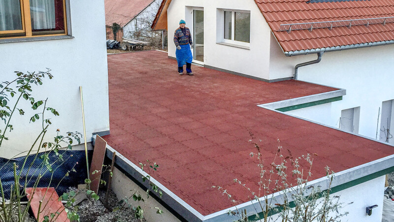 Zwischen zwei Einfamilienhäusern liegt ein ein Anbau mit Flachdach. Dessen Flachdach wird als Terrasse gestaltet und so für die Bewohner der angrenzenden Häuser nutzbar gemacht. Die Dachterrasse besteht aus roten Terrassenplatten von WARCO.