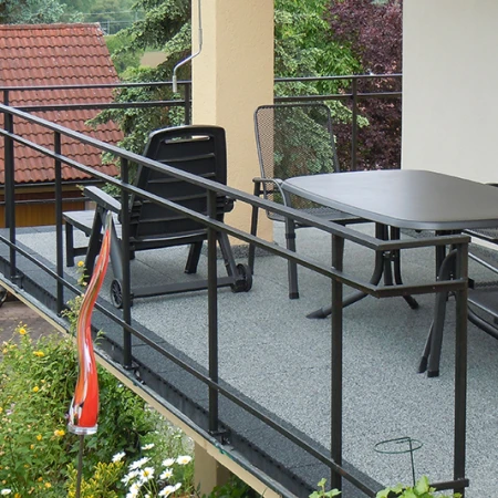Auf dem Bild sieht man einen geräumigen Balkon, dessen Boden mit grauen WARCO-Bodenplatten ausgelegt ist. Ein schwarzes Metallgeländer umrahmt die Fläche und bietet Sicherheit. Dazu passen der großzügige Tisch und die Stühle, die zum geselligen Beisammensein einladen. Der elastische, rutschfeste Belag ist witterungsbeständig und pflegeleicht, wodurch dieser Außenbereich dauerhaft geschützt ist und zum Entspannen einlädt.