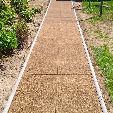 Une allée droite de jardin en dalles carrées de caoutchouc avec bordure latérale traverse le massif. Les dalles brunes en granulés de caoutchouc offrent une surface antidérapante, résistante aux intempéries et facile d’entretien. L’allée assure une marche sûre et propre par tous les temps et sépare nettement les parterres de la pelouse. Idéal pour jardins, cours et accès.