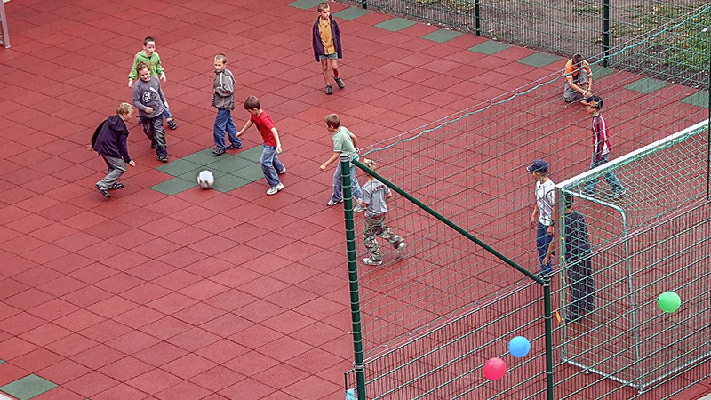 Des enfants jouent au football sur le nouveau terrain de foot de l'école, réalisé avec des dalles sportives rouges et vertes de WARCO.