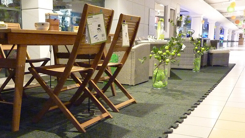 Im Einkaufszentrum wurde ein Teil der gekachelten Allee für den Sonderverkauf von Gartenmöbeln abgetrennt. Die Gartenmöbel stehen auf grünen Messeboden Klickfliesen.