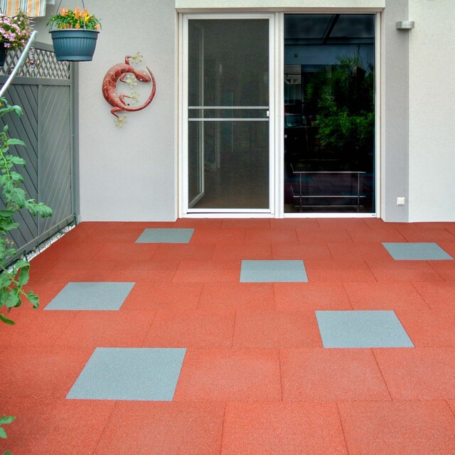 Die Terrasse nimmt die gesamte Hausbreite vor einem modernen Reihenhaus ein. Die rote Farbe der Terrassenplatten bildet einen wohltuenden Kontrast zu den Grautönen des Hauses.