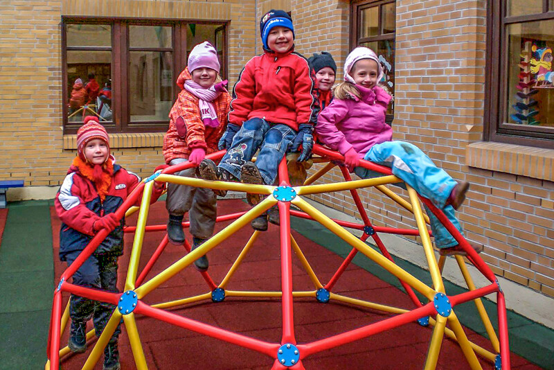 Auf einem röhrenförmigen Klettergerüst mit mehreren Oberflächen sitzend, lächeln spielende Kinder den Betrachter an. Unter dem Spielgerüst, das vor einer Hausecke aufgebaut ist, befinden sich rote und grüne Fallschutzplatten von WARCO.